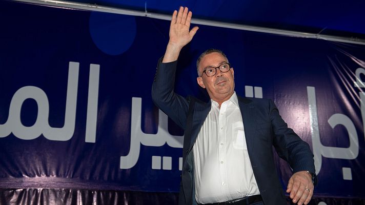 Primer plano de Fouzi Lekjaa saludando, con traje oscuro, camisa blanca y gafas. Fondo con pancarta árabe y detalle azul.