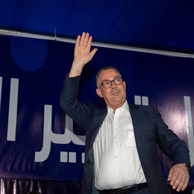 Primer plano de Fouzi Lekjaa saludando, con traje oscuro, camisa blanca y gafas. Fondo con pancarta árabe y detalle azul.