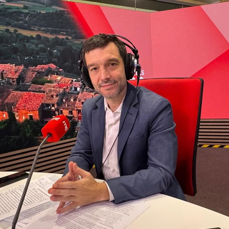 Pablo Bustinduy en estudio de radio con micrófono "rne", chaqueta azul marino, camisa blanca, folios y fondo de paisaje rural y panel rojo con "S".
