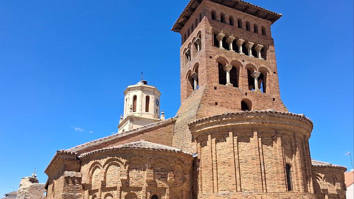 Edificio religioso mudéjar con torre campanario de ladrillo y otra torre octogonal con reloj y cúpula, bajo cielo azul.