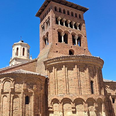 Edificio religioso mudéjar con torre campanario de ladrillo y otra torre octogonal con reloj y cúpula, bajo cielo azul.