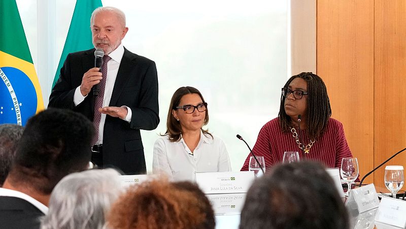 Lula da Silva habla en un evento oficial, acompañado por Janja Lula da Silva y otra mujer. La bandera de Brasil ondea al fondo.