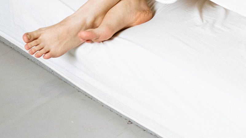 Primer plano de dos pies descalzos sobre un colch�n blanco arrugado, con suelo gris claro debajo. Ambiente tranquilo y de descanso.