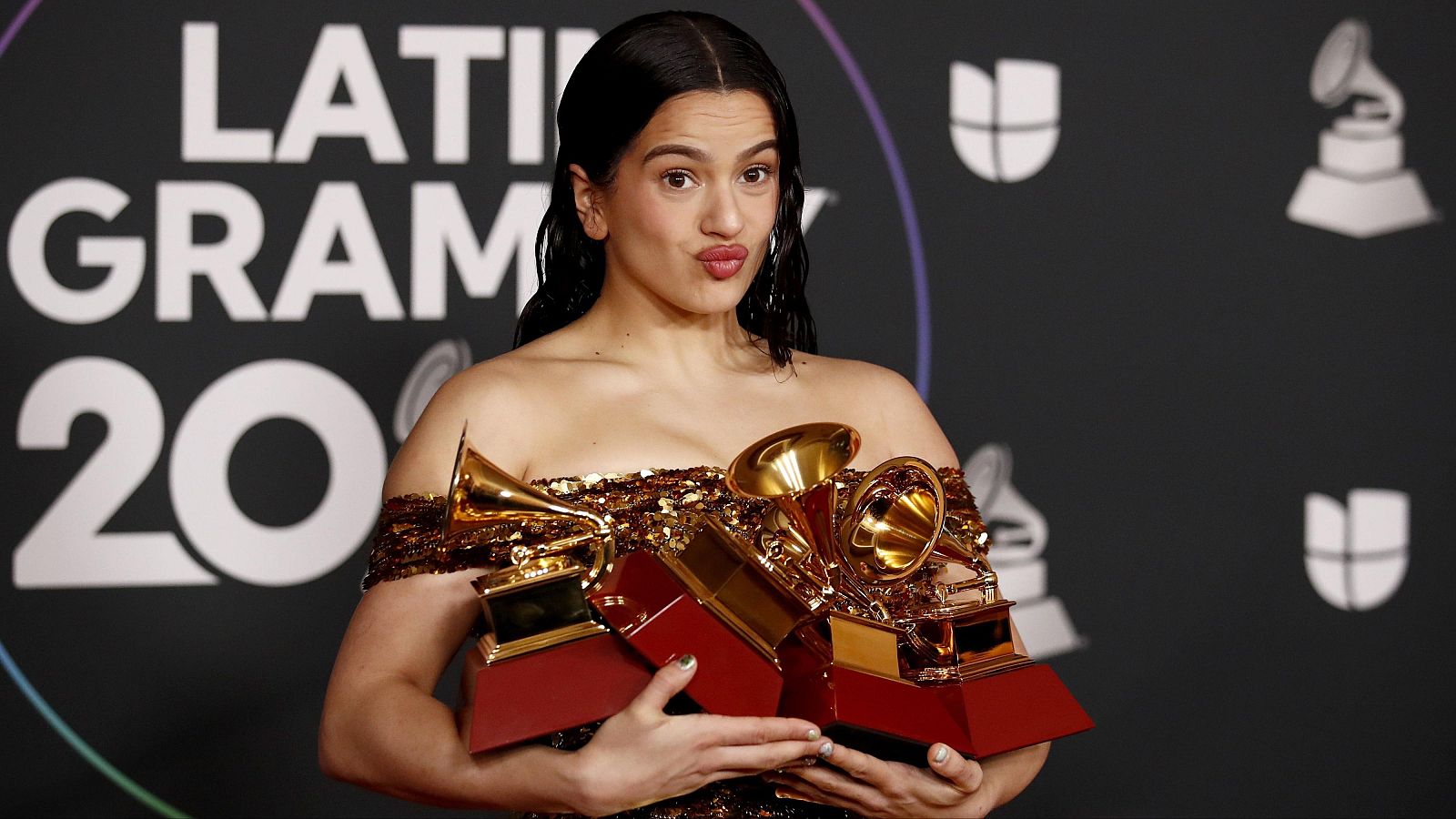 Rosal�a, con top dorado y cabello recogido, posa con tres trofeos dorados de gram�fono en un fondo negro con "LATI GRAM 20" y logos de Grammy/Univision.