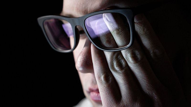 Persona con gafas oscuras, mano cubriendo ojo derecho. Reflejos viol�ceos y azulados en las lentes. Fondo oscuro.