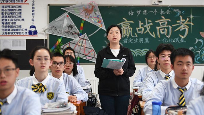 Profesora dirige clase en aula china. Estudiantes con uniformes y corbatas a rayas asisten. Pizarra muestra lemas para el Gaokao y hábitos de estudio.