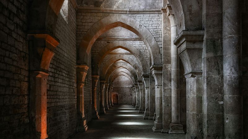 Pasillo arquitect�nico antiguo con arcos apuntados, pilares de piedra y b�vedas. Iluminaci�n tenue y dram�tica con luz c�lida anaranjada en el lado izquierdo. Profundidad y misterio.
