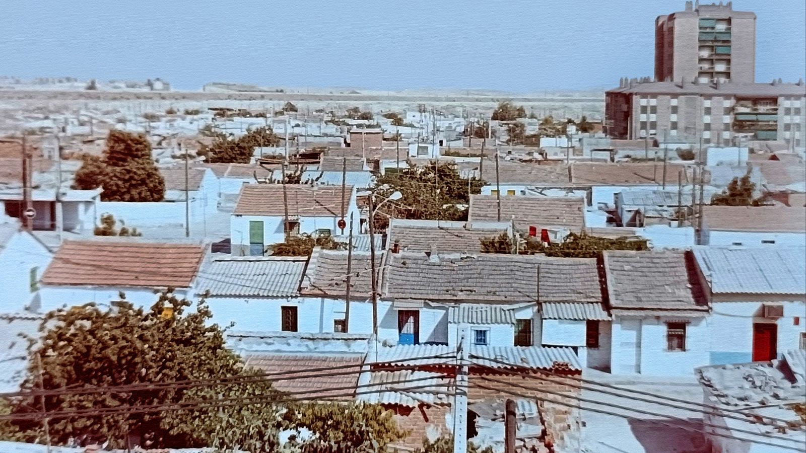 Vista aérea de un barrio residencial con casas blancas y tejados rojizos, postes de luz y un edificio moderno al fondo. El cielo está despejado y el ambiente es soleado.