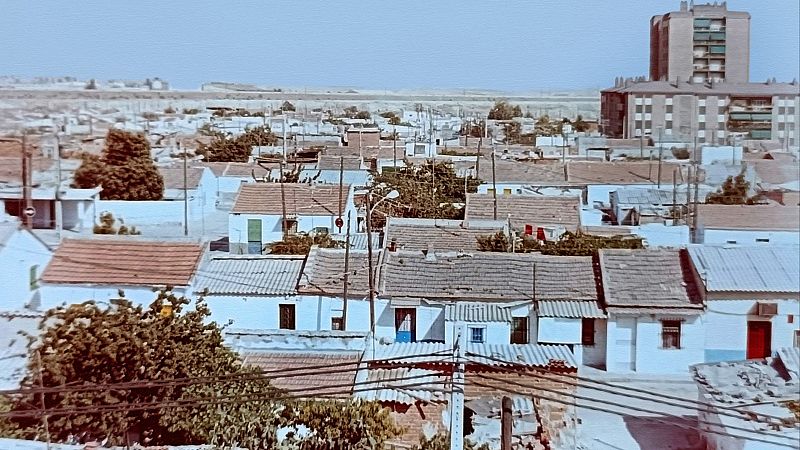 Vista aérea de un barrio residencial con casas blancas y tejados rojizos, postes de luz y un edificio moderno al fondo. El cielo está despejado y el ambiente es soleado.