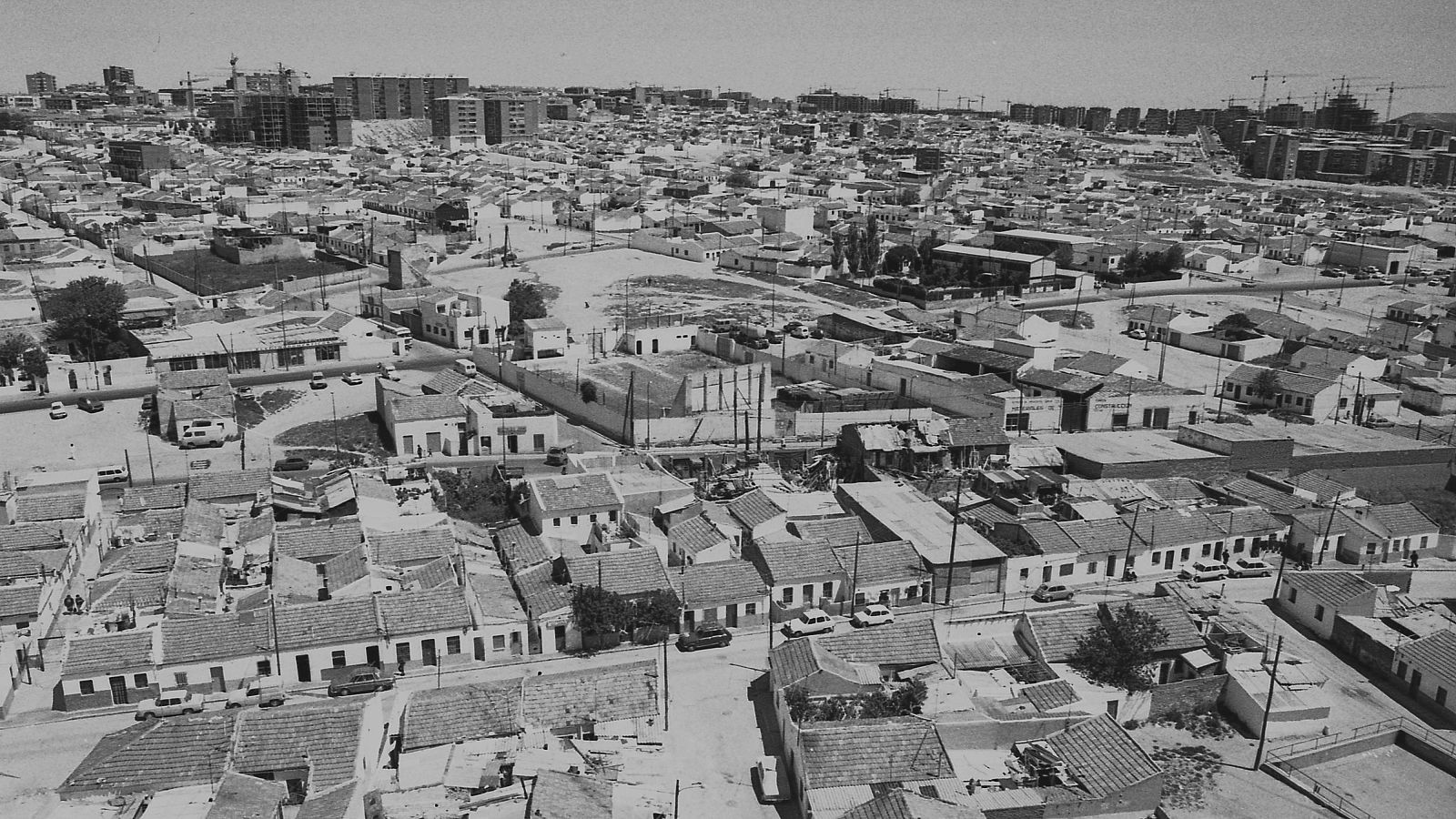 Vista aérea en blanco y negro de una zona urbana con casas bajas densas y calles estrechas, contrastando con edificios altos y grúas de construcción al fondo, indicando desarrollo urbano. Palomeras Bajas, Madrid.