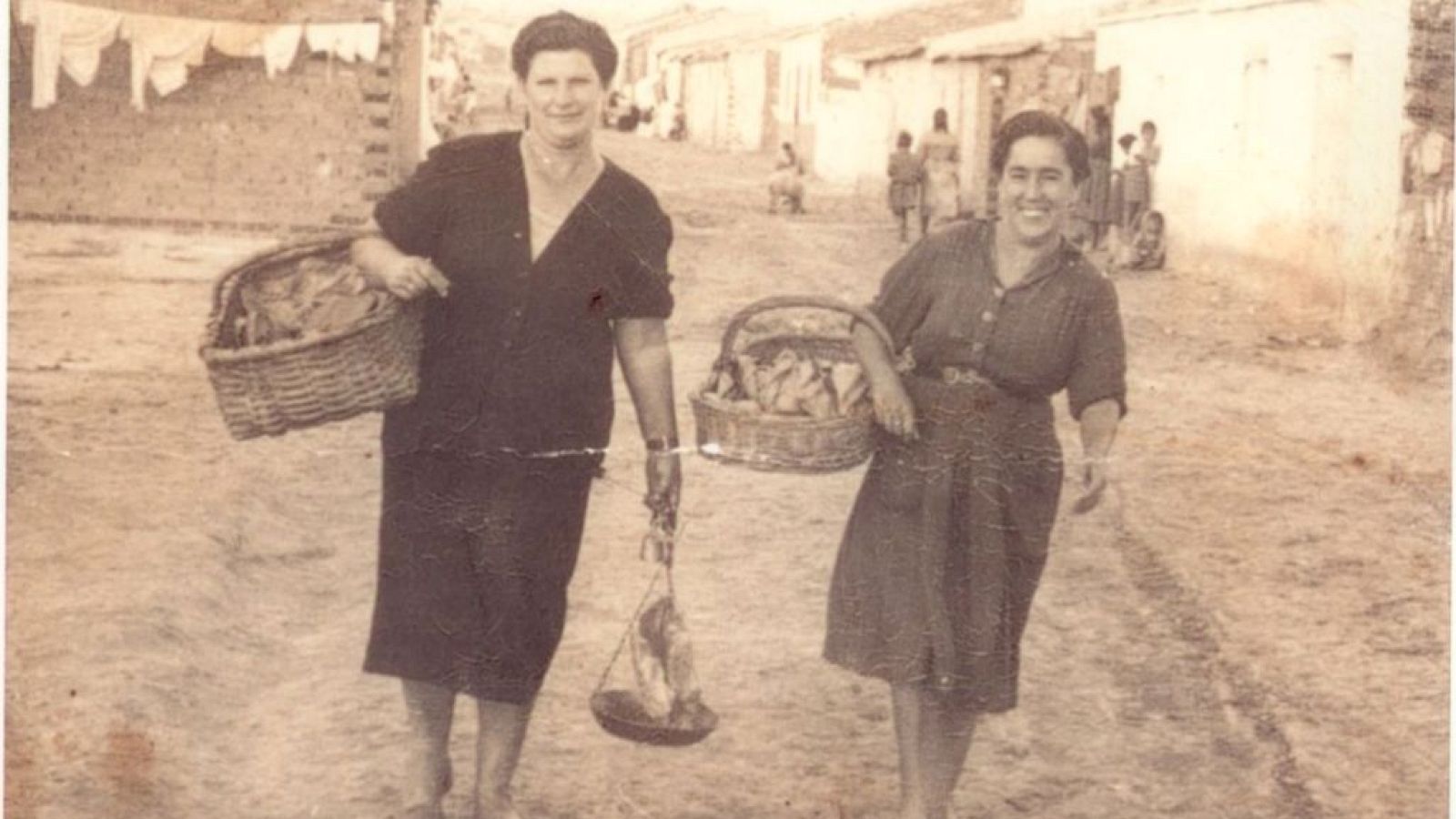 Dos mujeres con cestas y una balanza romana caminan por una calle de tierra, rodeadas de edificaciones humildes y ropa tendida, en una escena de la vida cotidiana de mediados del siglo XX.
