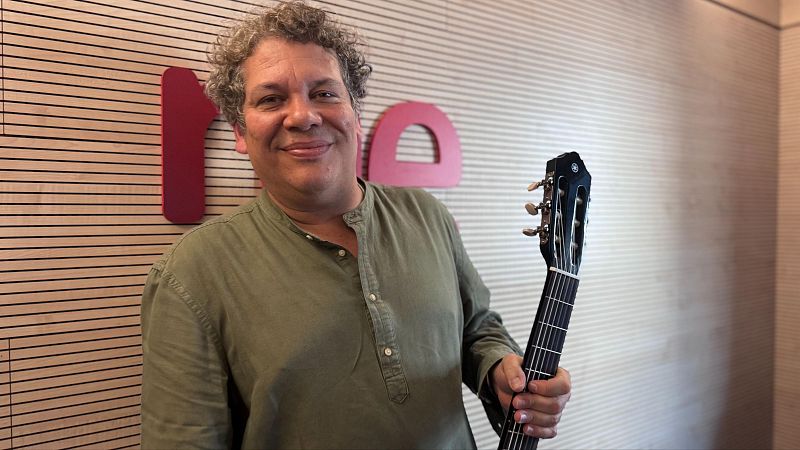 Primer plano del músico colombiano Chabuco sonriendo, sosteniendo una guitarra. Viste camisa verde oliva y su cabello es grisáceo y rizado. Fondo con letras rojas y patrón de madera.