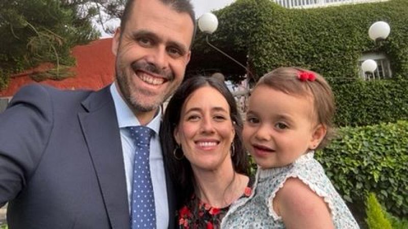 Retrato familiar al aire libre: hombre con traje azul, mujer con vestido estampado y niña sonriente, frente a edificio cubierto de hiedra y lámparas esféricas.