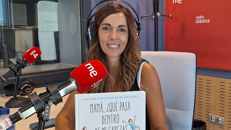 Elena Sanz, con auriculares y sosteniendo su libro "MAMÁ, ¿QUÉ PASA DENTRO DE MI CABEZA?", en un estudio de radio con micrófonos de RNE y Ràdio 4.