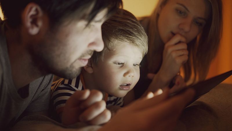 Familia reunida en la oscuridad, iluminada por la pantalla de una tablet, mostrando concentración y asombro.