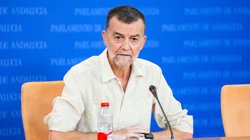 Antonio Maíllo, con cabello y barba canosa, viste camisa clara y mira a la cámara en el Parlamento de Andalucía, con micrófono y botella de agua.