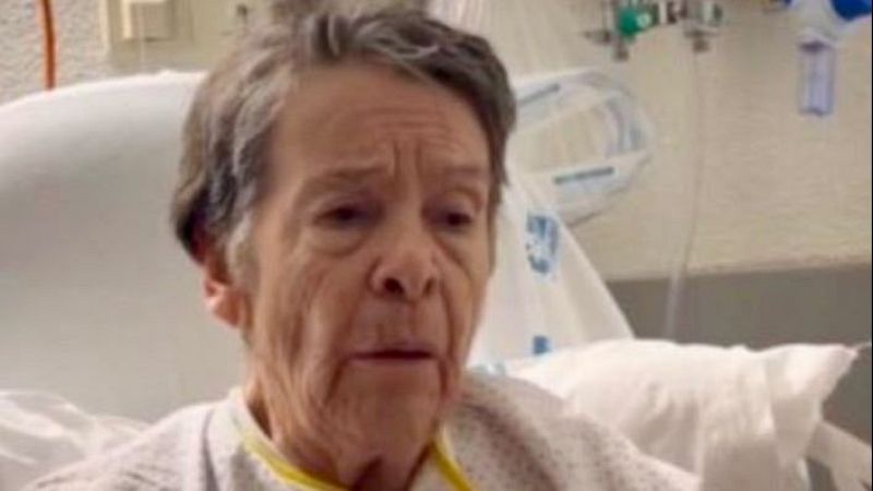 Mari Carmen, mujer mayor con bata de hospital, rostro cansado y arrugado, en un entorno m�dico con equipo visible al fondo.