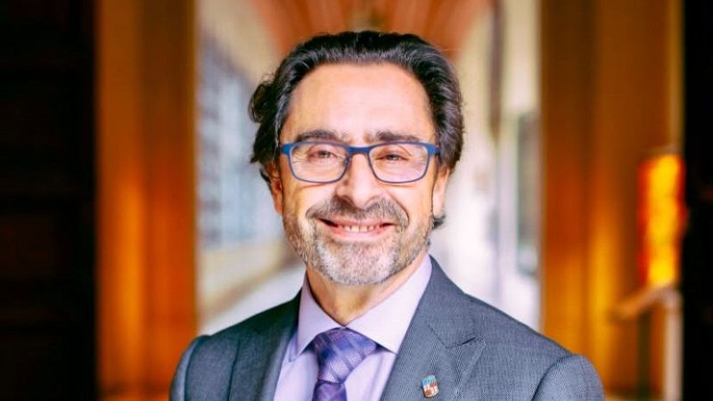 Retrato de primer plano de Joan Gu�rdia, hombre de mediana edad a mayor, sonriendo a la c�mara con gafas azules y pin her�ldico en chaqueta gris.