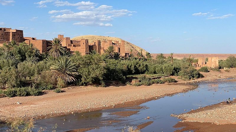 Impresionante ksar de adobe sobre una colina, rodeado de un oasis verde con palmeras junto a un r�o, bajo un cielo azul y soleado. Paisaje des�rtico con estructuras adicionales al fondo.