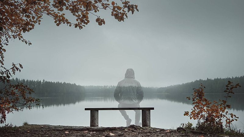 Figura translúcida sentada en banco de madera frente a lago brumoso y bosque otoñal. Atmósfera de introspección y misterio.