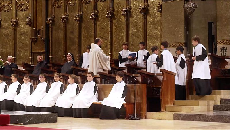 Monje dirige a coro infantil en bas�lica. Ni�os con t�nicas negras y blancas, algunos con partituras, en siller�a de coro de madera oscura. Adultos observan.