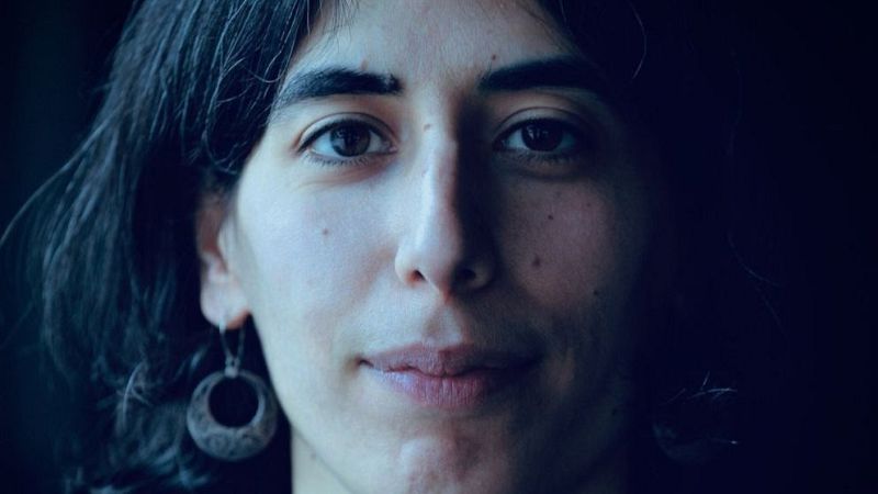 Retrato en primer plano de la compositora Nuria Gim�nez Comas, con expresi�n serena, cabello oscuro y un pendiente de plata circular, bajo una iluminaci�n de tonos fr�os.
