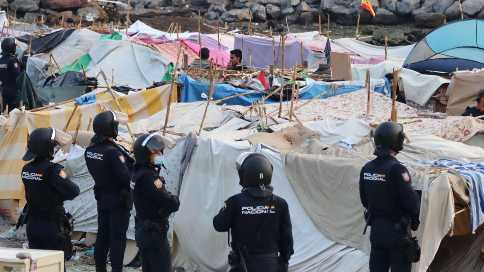 Agentes de la Policía Nacional observan un campamento de tiendas improvisadas en un terreno rocoso, con una bandera de España al fondo.