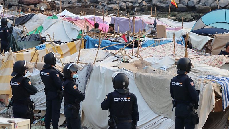 Agentes de la Policía Nacional observan un campamento de tiendas improvisadas en un terreno rocoso, con una bandera de España al fondo.