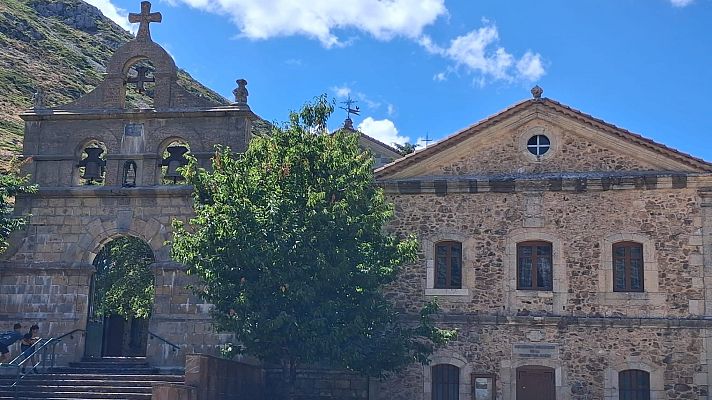 Complejo de piedra con campanario y ermita en entorno montañoso, bajo cielo azul con nubes. Figuras humanas en escaleras.