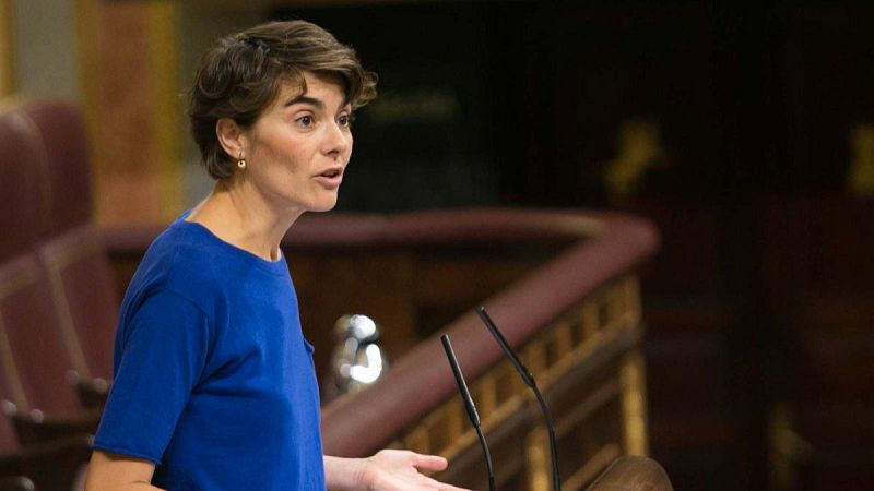 Yolanda Díaz, de perfil, con la boca abierta y mano izquierda levantada, en un hemiciclo. Dos micrófonos negros en primer plano.