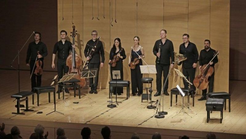 Ocho m�sicos en un escenario de concierto, vestidos de negro, tocan diversos instrumentos como violines, contrabajo, fagot, clarinete, trompa y violonchelo. El p�blico aplaude al final.