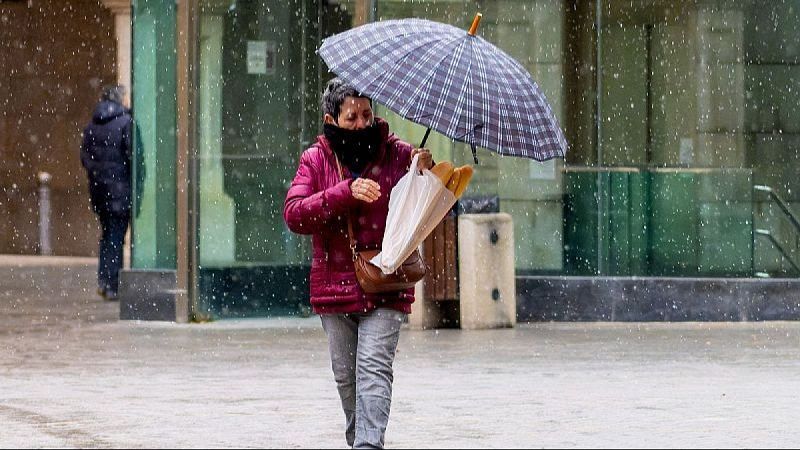 Mujer con paraguas a cuadros y bolsa de pan camina bajo intensa lluvia en entorno urbano con edificios modernos y suelo mojado.