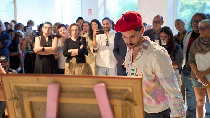 Hombre con boina roja y camisa manchada de pintura, frente a un caballete, en un evento cultural con público atento.