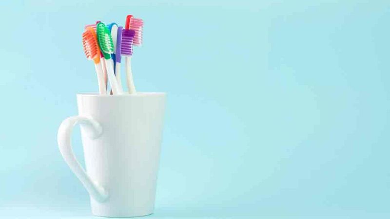 Taza blanca con cinco cepillos de dientes de cerdas de colores (naranja, verde, azul, morado, rosa) en su interior, sobre fondo azul claro. Ideal para lavarse los dientes.