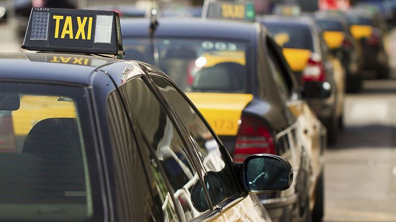 Primer plano de un taxi oscuro con cartel "TAXI" amarillo. Detr�s, otros taxis con franja trasera amarilla. Iluminaci�n diurna.