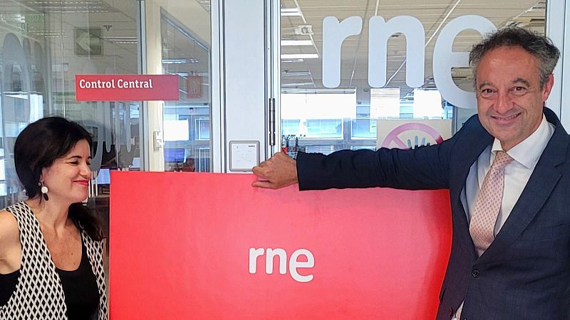 José Manuel Pérez Tornero y María Eizaguirre sonríen en RNE. Él, con traje azul, apoya la mano en un panel rojo con el logo 'rne'. Ella, con chaleco geométrico, mira hacia él.