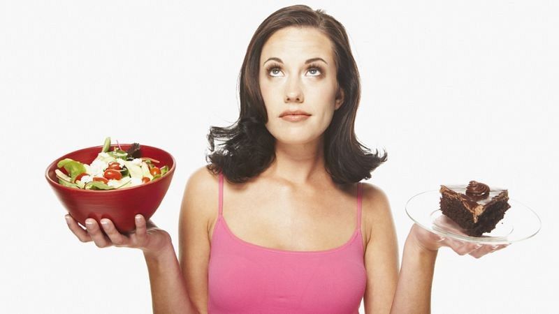 Mujer pensativa elige entre ensalada y tarta de chocolate, con mirada hacia arriba y boca cerrada. Sostiene ensalada en mano izquierda y tarta en mano derecha.
