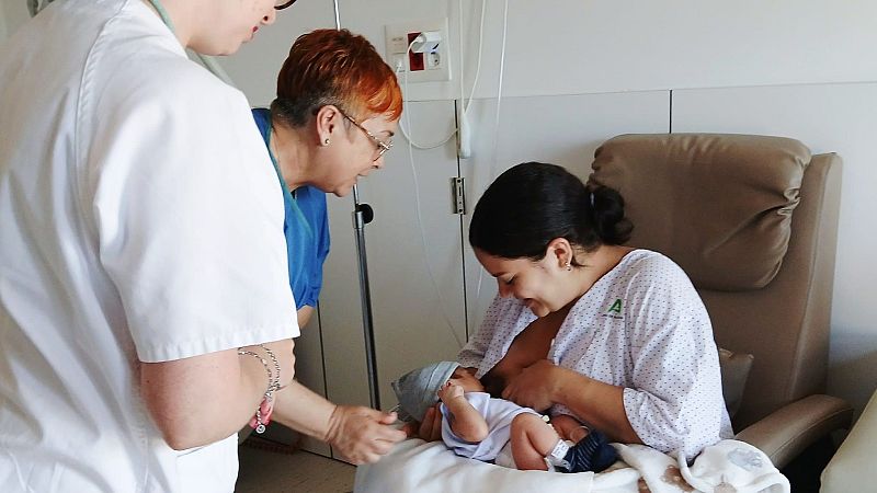 Una madre con bata de la Junta de Andalucía amamanta a su bebé en un hospital, acompañada por dos sanitarias que la observan con afecto en una luminosa habitación clínica.