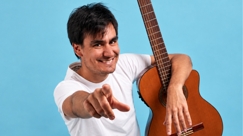 Pau de Ponts, m�sico y presentador, sonr�e y apunta a la c�mara mientras sostiene una guitarra ac�stica. Fondo azul claro.