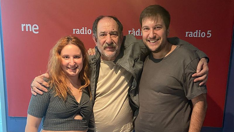Carlos Latre posa sonriente con una mujer y un hombre en un estudio de radio de RNE. Se distinguen los logos "rne" y "radio 5" en la pared roja.