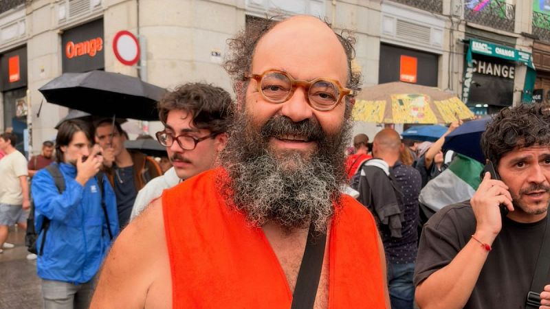 El Gran Wyoming, con barba y gafas, sonríe a cámara en una calle urbana lluviosa, rodeado de gente con paraguas y letreros de "Orange" y "CHANGE".