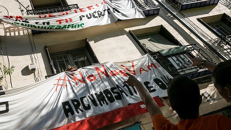 Protesta social con pancartas "NO TE VAYAS DE TU PUERTA NOS QUEDAMOS" y "NO NOS VAMOS ARCUMOSATIE QUE". Dos personas señalan los mensajes.