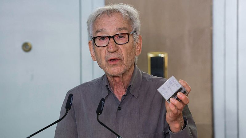 Primer plano de José Luis Cuerda, director de cine, con gafas y cabello canoso, hablando ante micrófonos y mostrando un objeto táctil con cuadrícula.