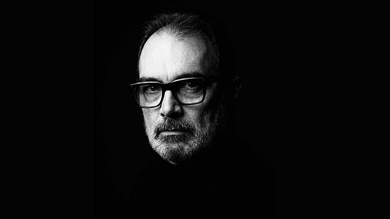 Retrato en blanco y negro de Joan Valent, compositor y m�sico espa�ol, con gafas de montura gruesa y barba entrecana, iluminado dram�ticamente sobre fondo negro.
