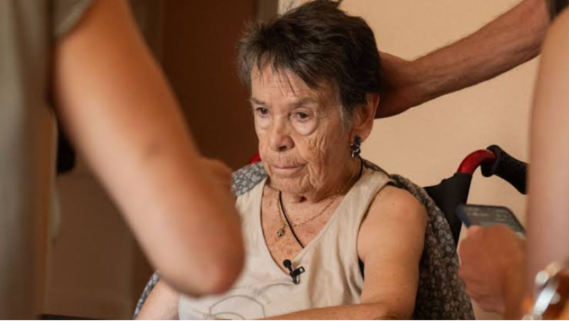 Mari Carmen Mart�nez-Villase�or Barrasa, con micr�fono, en una grabaci�n, asistida por otros. Posiblemente en silla de ruedas, con manta y expresi�n pensativa.
