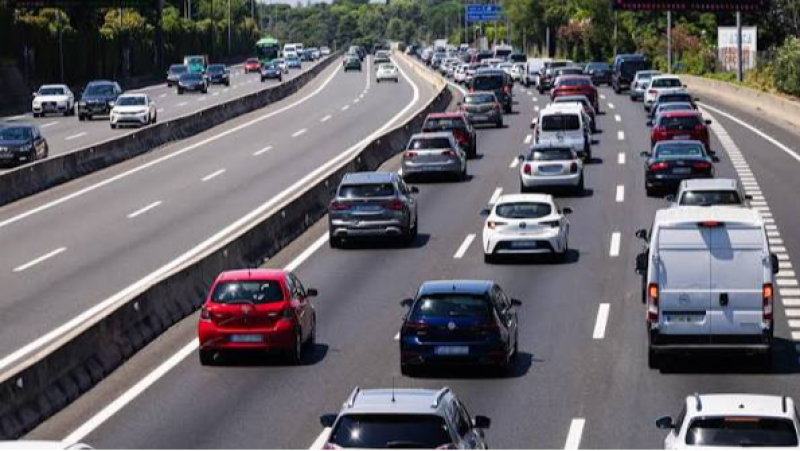 Vista a�rea de autopista con tr�fico denso. Coches rojos, azules, grises y blancos, junto a furgonetas, circulan en carriles separados por mediana de hormig�n. Entorno natural con vegetaci�n.