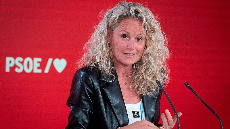 Montserrat M�nguez, pol�tica del PSOE, habla en un evento con micr�fonos. Viste chaqueta negra y camiseta blanca. Fondo rojo con logo del PSOE.