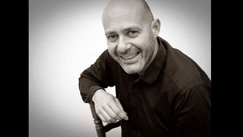 Retrato en blanco y negro de Curro Pi�ana, cantaor de flamenco, sonriendo y mirando a c�mara. Apoya su brazo en una silla de madera sobre fondo liso.