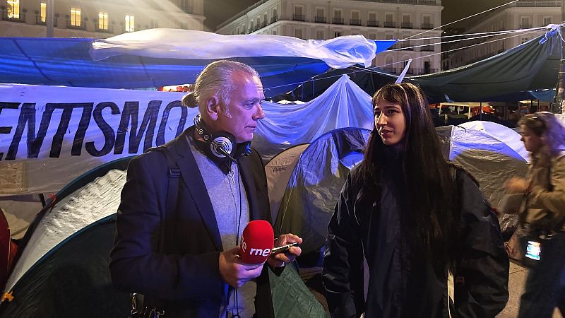 Periodista Manolo H. H. de RNE entrevista a una joven en un campamento de protesta nocturno, con tiendas y una pancarta visible. Iluminación azulada.