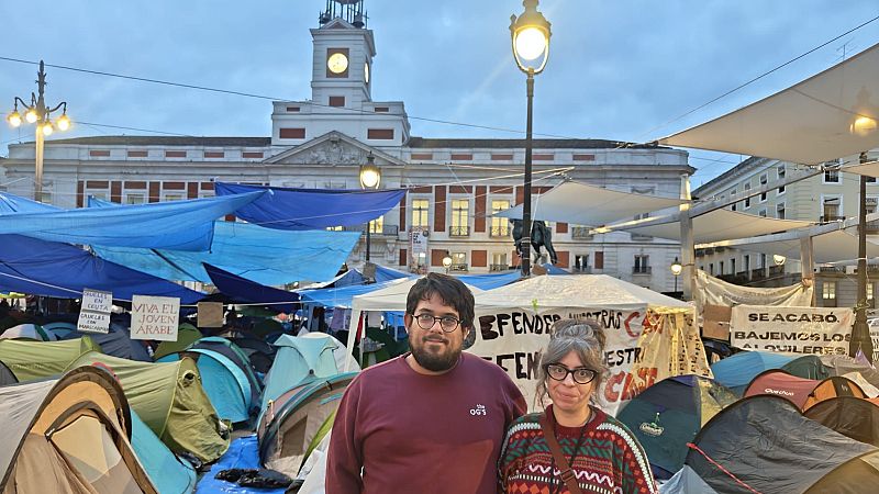 Campamento de protesta en la Puerta del Sol, Madrid. Hombre y mujer con gafas en primer plano, rodeados de tiendas y pancartas reivindicativas. Edificio de Correos al fondo.